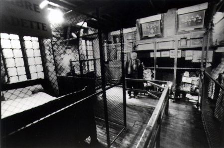 Photo de la Chambre de Bernadette à Lourdes. À gauche, on remarque le lit, le traversin et sur ce dernier, l'écriteau indiquant que c’était le sien. À droite, sous les portraits de famille, la grande cheminée qu’elle pouvait voir facilement en s’endormant.